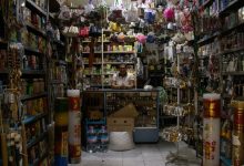 Comerciante atrás do balcão de uma pequena loja repleta de produtos diversos.