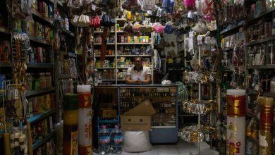 Comerciante atrás do balcão de uma pequena loja repleta de produtos diversos.