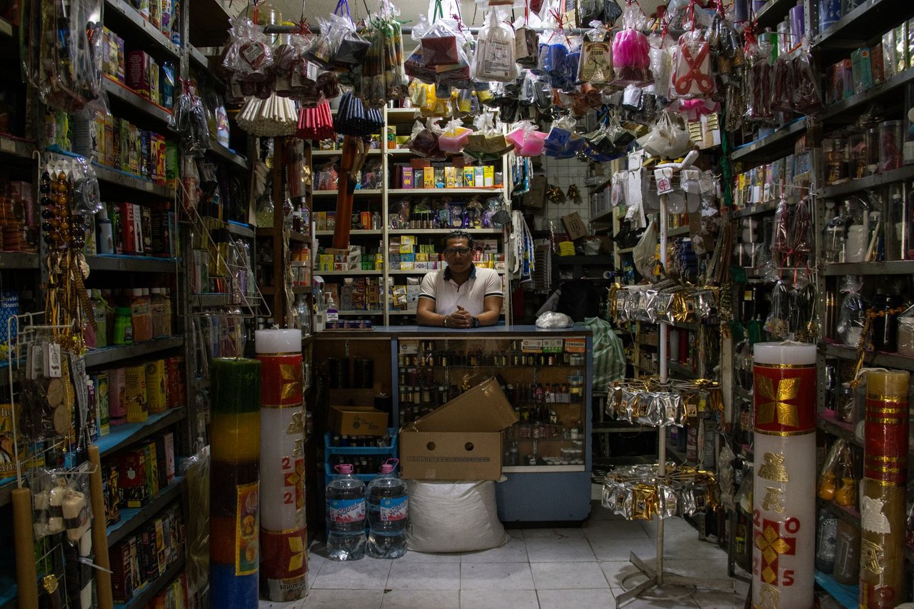 Comerciante atrás do balcão de uma pequena loja repleta de produtos diversos.