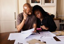 Casal sentado à mesa organiza documentos financeiros enquanto consulta um celular.