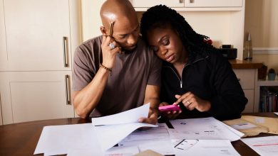 Casal sentado à mesa organiza documentos financeiros enquanto consulta um celular.