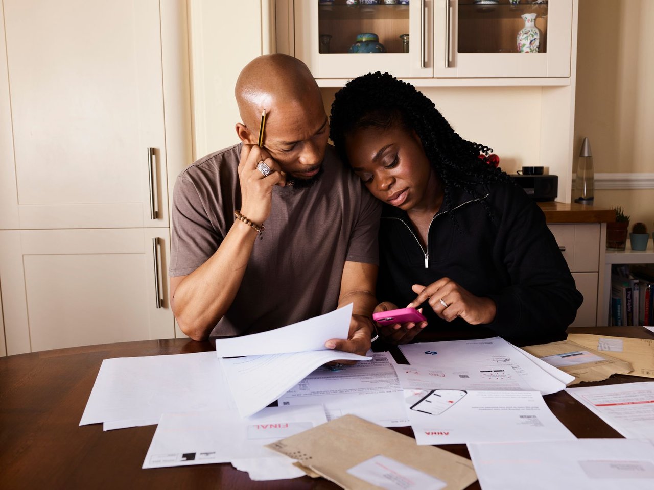Casal sentado à mesa organiza documentos financeiros enquanto consulta um celular.