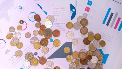 Moedas de diferentes valores sobre documentos financeiros com gráficos e linhas de mercado ao fundo.
