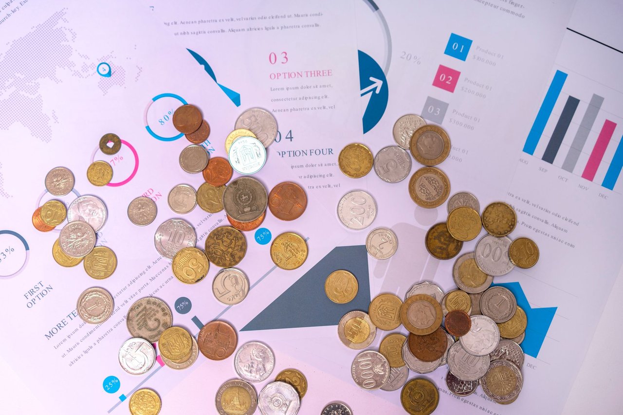 Moedas de diferentes valores sobre documentos financeiros com gráficos e linhas de mercado ao fundo.